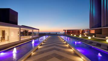 Un espace piscine moderne dans un hôtel, avec un motif en chevron, des jeux d’eau et une vue sur la mer au crépuscule.