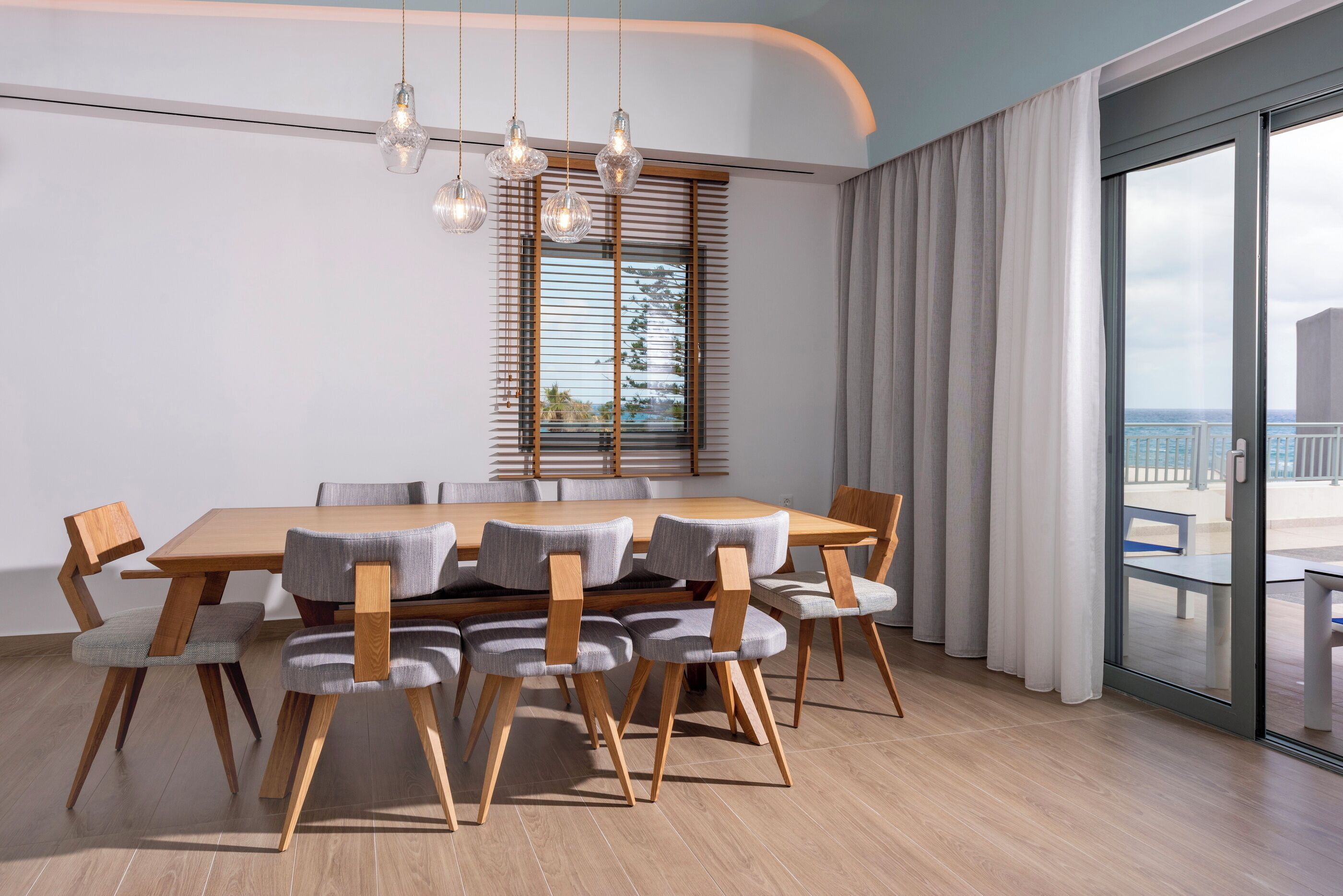 A modern dining area with a wooden table and chairs, a large window with blinds, and a view of the outdoors.