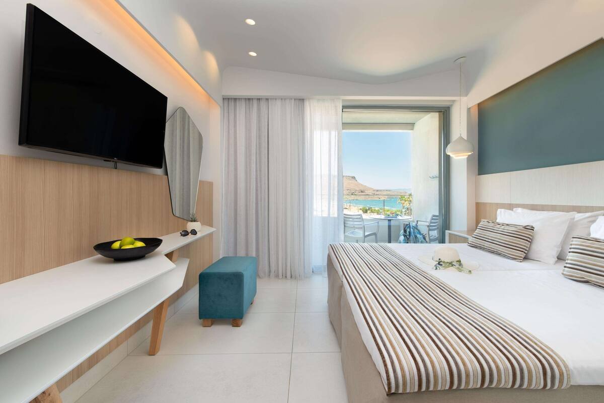 A modern hotel room with a flat-screen TV, a large bed with striped bedding, a small blue ottoman, a white bedside table with a bowl of fruit, and a view of the outdoors.
