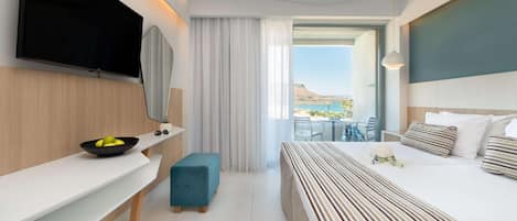 A modern hotel room with a flat-screen TV, a large bed with striped bedding, a small blue ottoman, a white bedside table with a bowl of fruit, and a view of the outdoors.