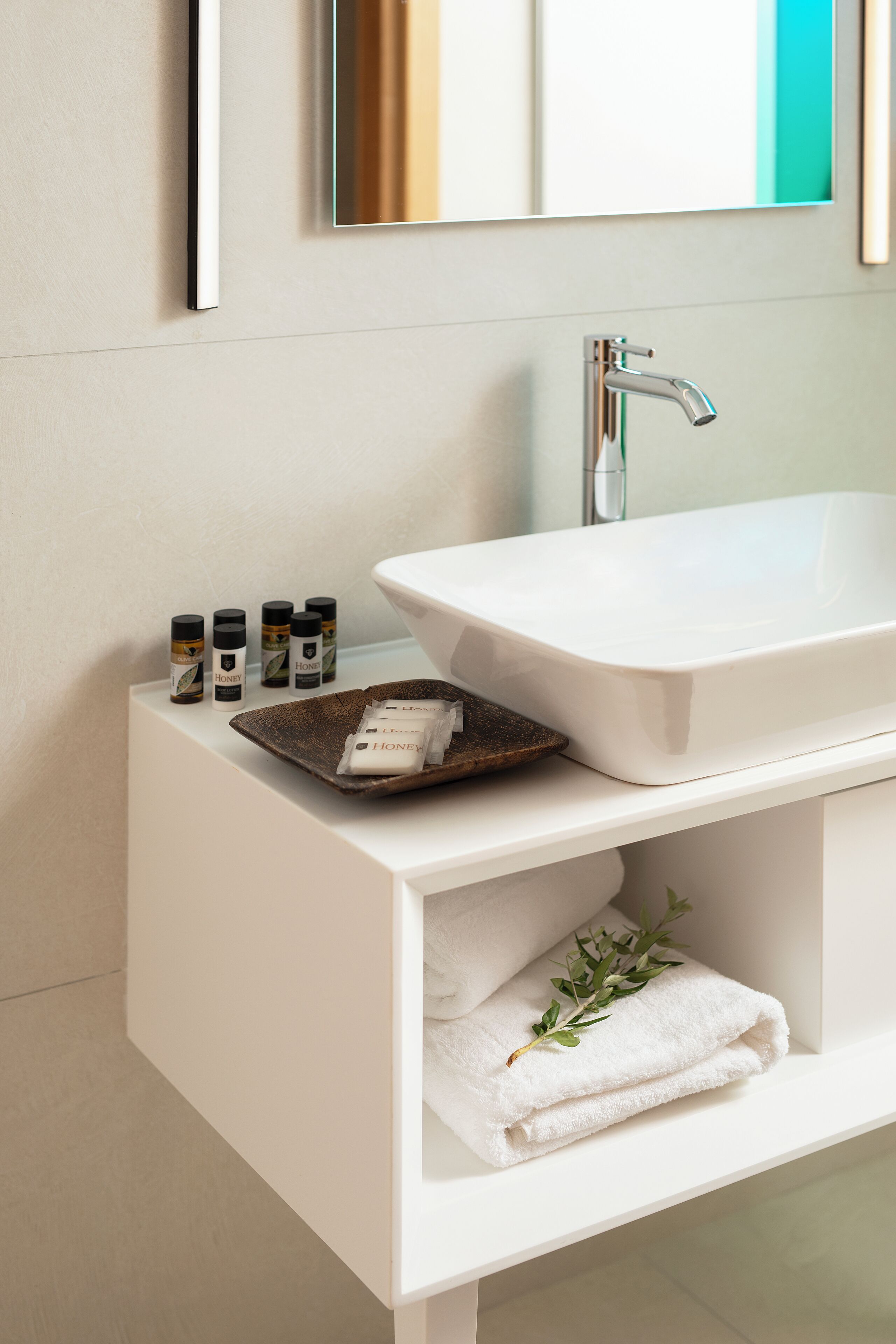Une salle de bain moderne avec un meuble-lavabo blanc, un lavabo rectangulaire et un miroir au-dessus. On y trouve quatre bouteilles de miel sur le meuble-lavabo, un plateau avec un savon, et une serviette blanche pliée ornée d’une feuille verte.