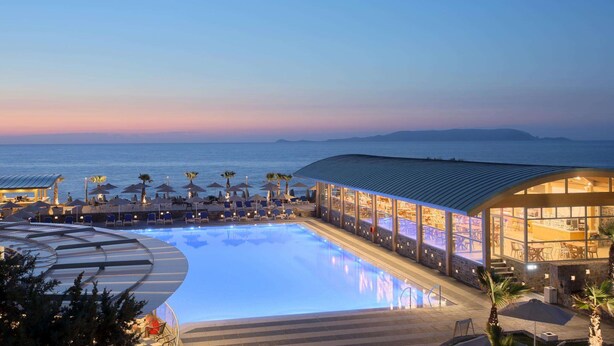 A swimming pool with lounge chairs and umbrellas, a bar area with large windows, and a view of the sea. - Arina Beach Resort - All inclusive (Hersonissos)