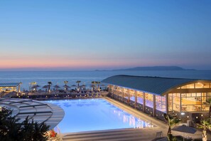 A swimming pool with lounge chairs and umbrellas, a bar area with large windows, and a view of the sea. - Arina Beach Resort - All inclusive (Hersonissos)