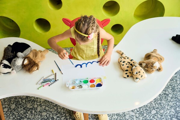 Niño dibujando en una mesa con materiales de arte, rodeado de peluches.