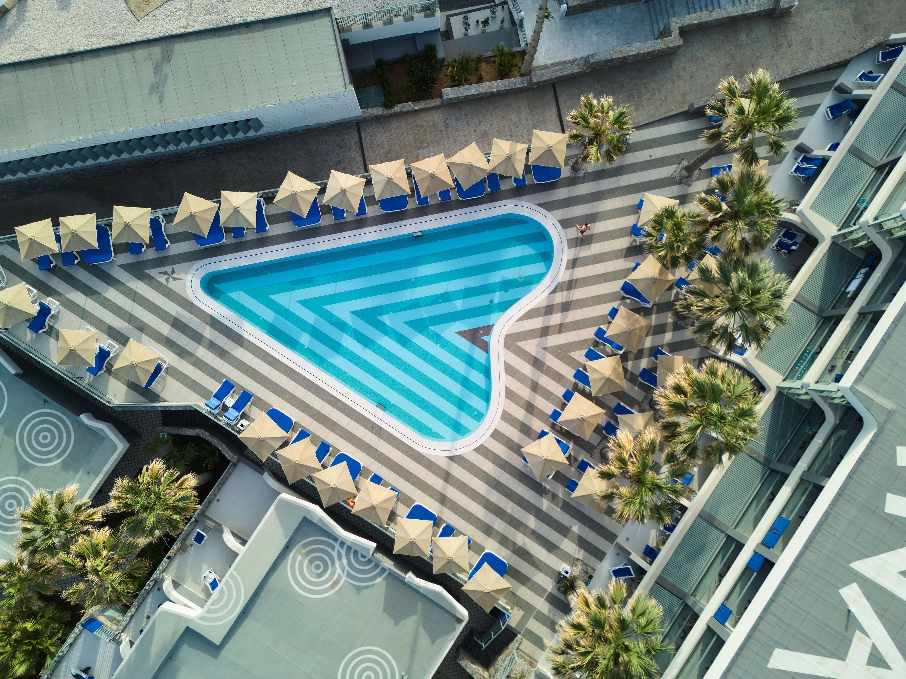 Ein Luftbild eines Pools mit blauen und weißen Streifen, umgeben von Liegestühlen und Sonnenschirmen, mit Palmen und einem modernen Gebäude im Hintergrund.