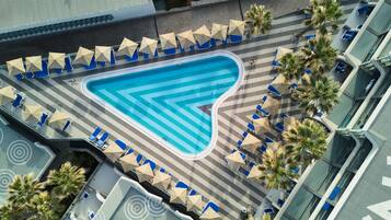 Ein Luftbild eines Pools mit blauen und weißen Streifen, umgeben von Liegestühlen und Sonnenschirmen, mit Palmen und einem modernen Gebäude im Hintergrund.