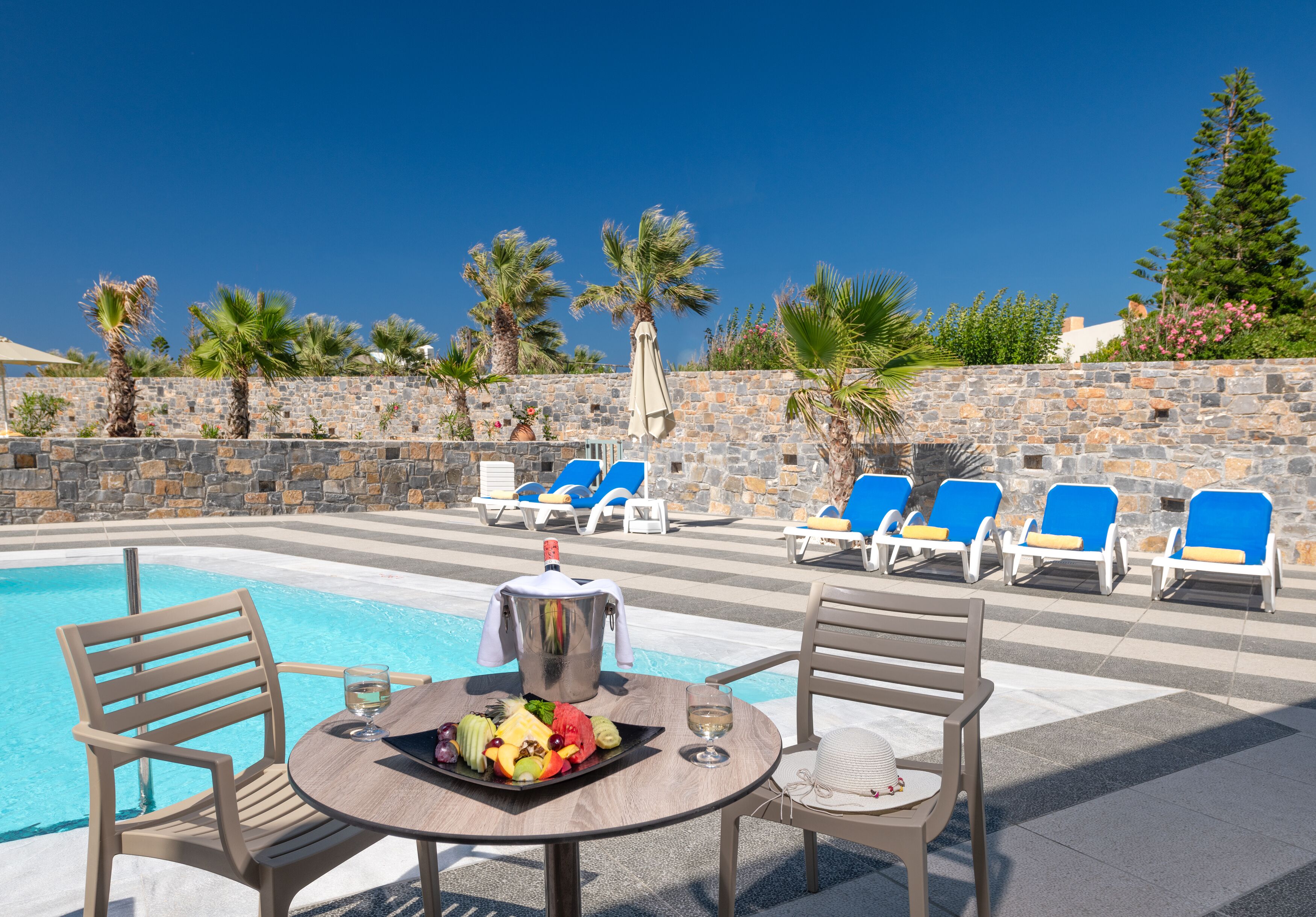 Zona de piscina con sillones, una mesa con frutas y una bebida, y un muro de piedra con palmeras.
