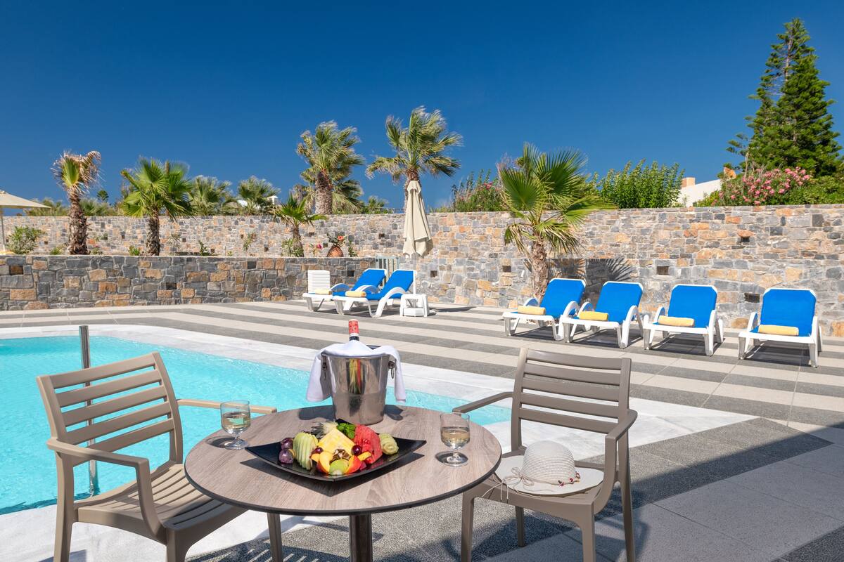 A poolside area with a table set for two, a fruit platter, and a pitcher. There are lounge chairs and a stone wall in the background.
