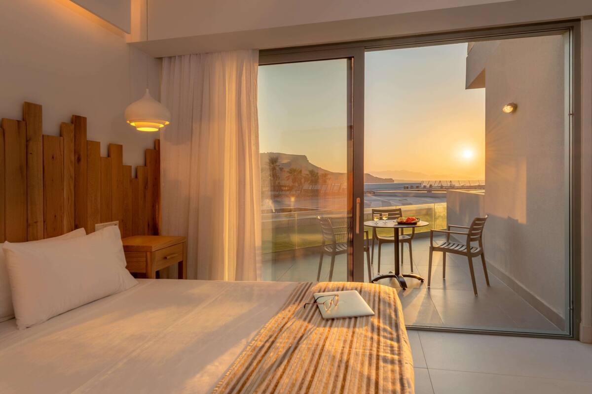 A hotel room with a bed, a laptop, a small table, and a balcony with a view of the sunset and a distant landscape.