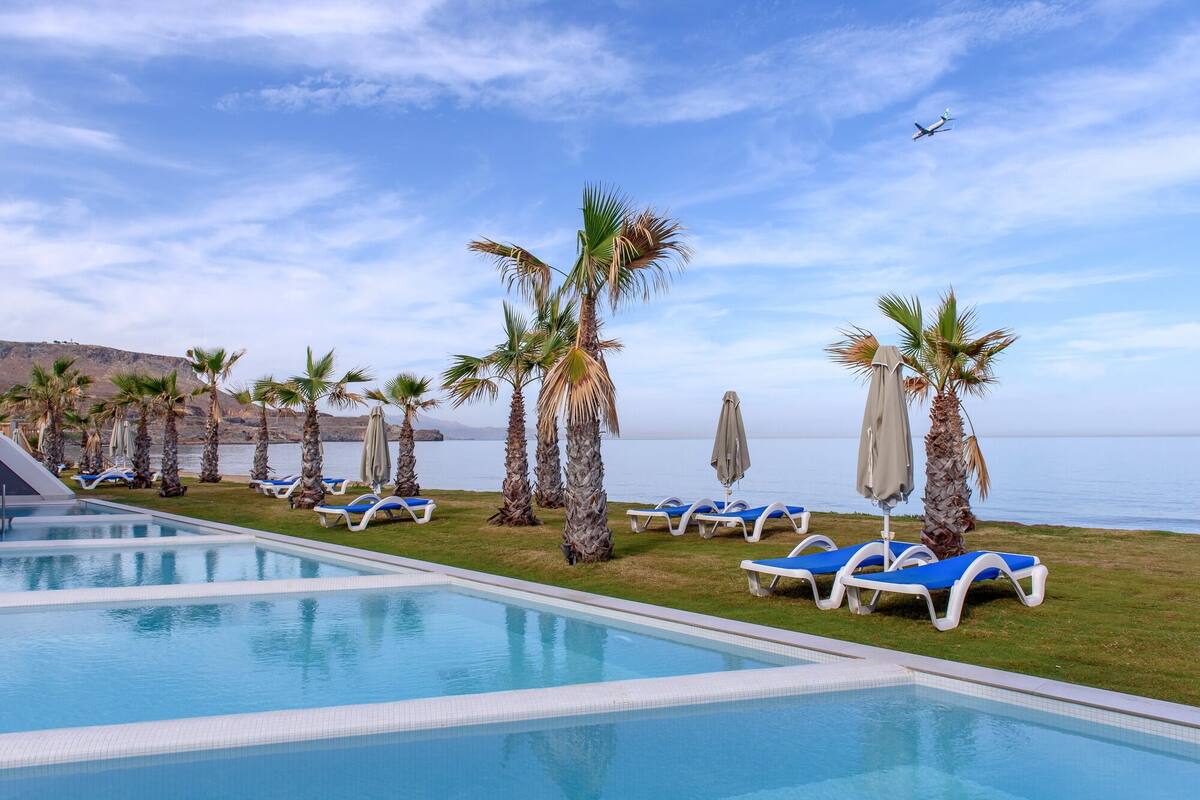 A pool area with lounge chairs and umbrellas, palm trees, and a view of the sea.