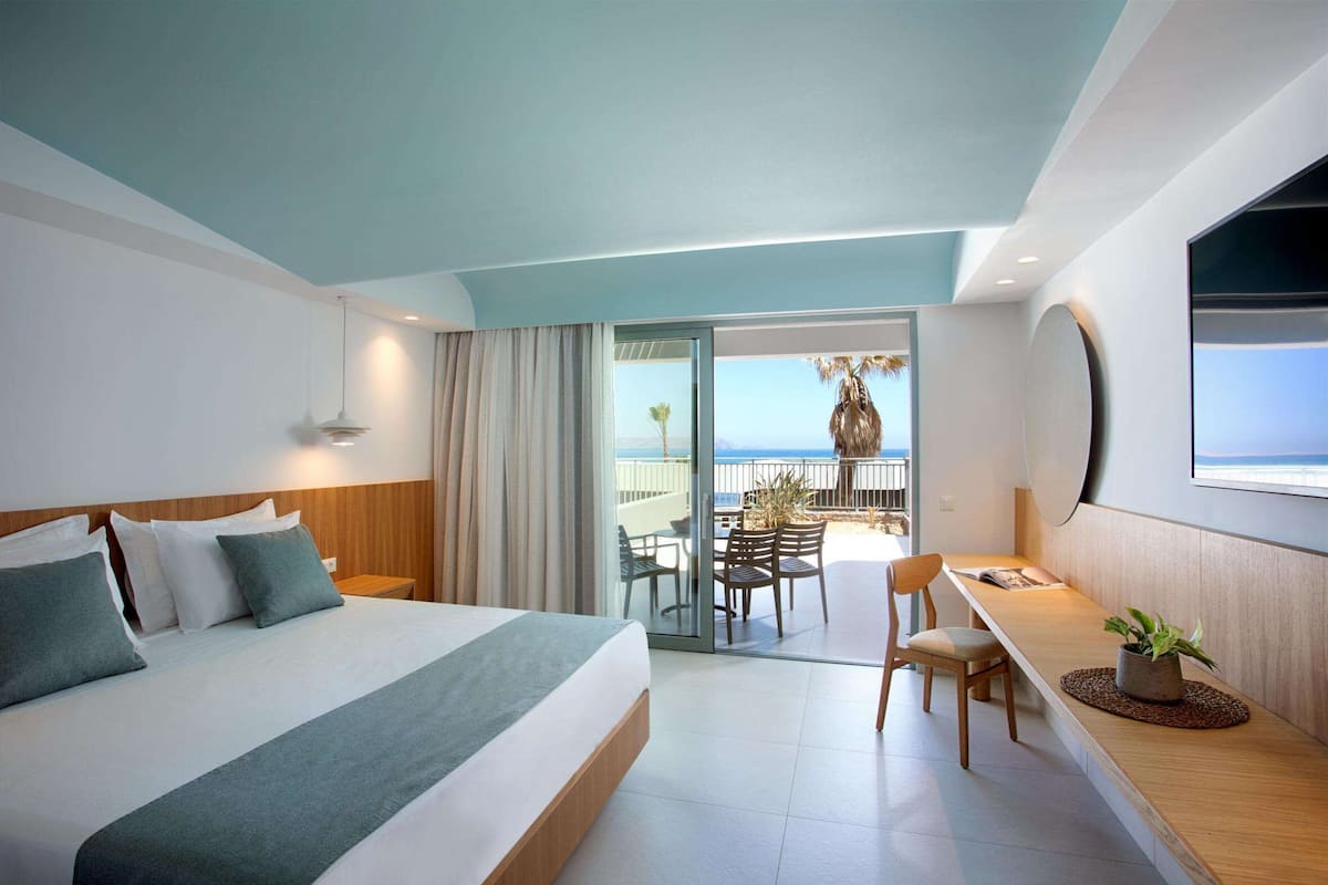 A modern hotel room with a large bed, a desk, and a balcony with a view of the sea.