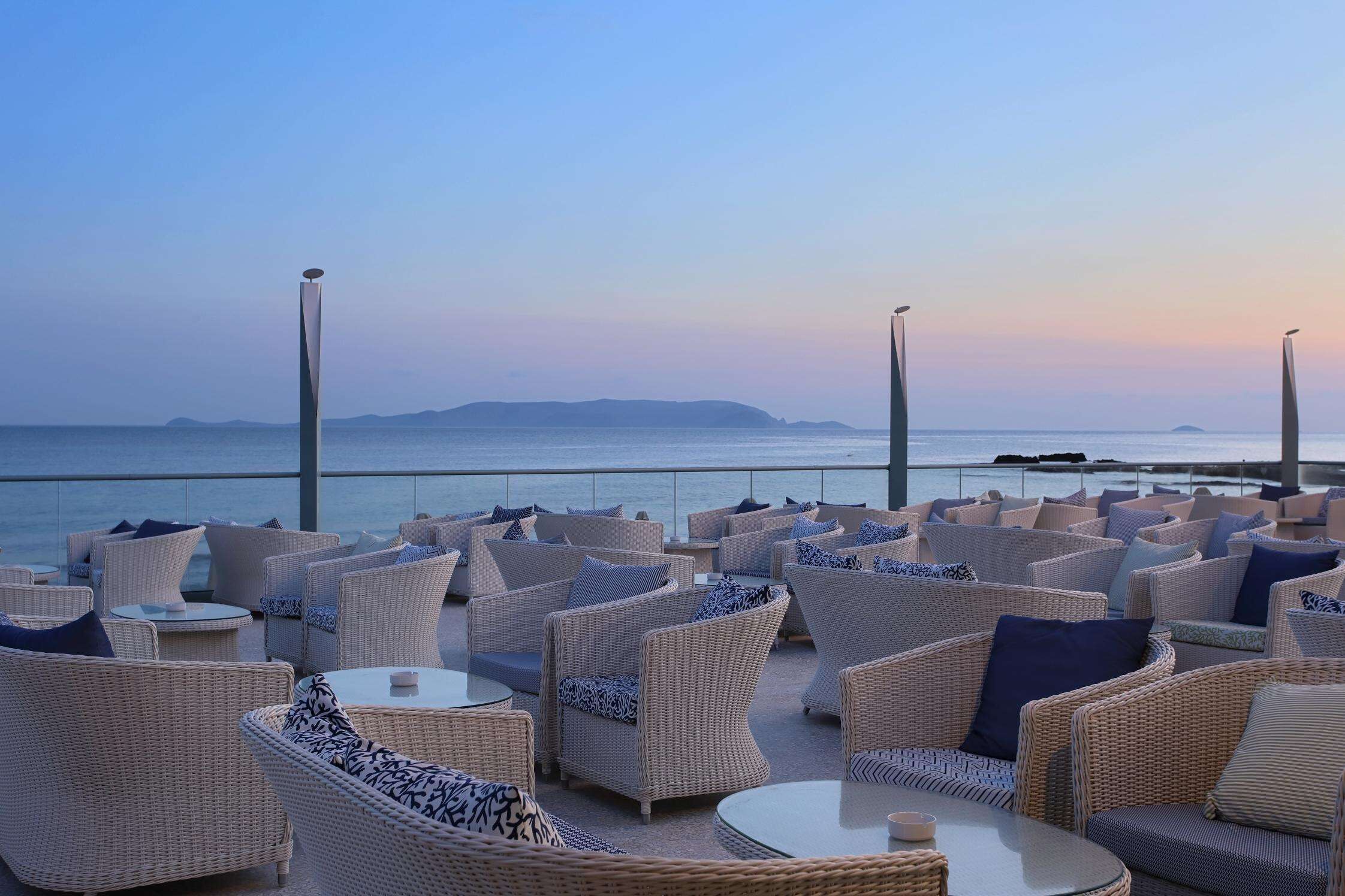 A rooftop terrace with wicker furniture and tables, overlooking a body of water and distant land.