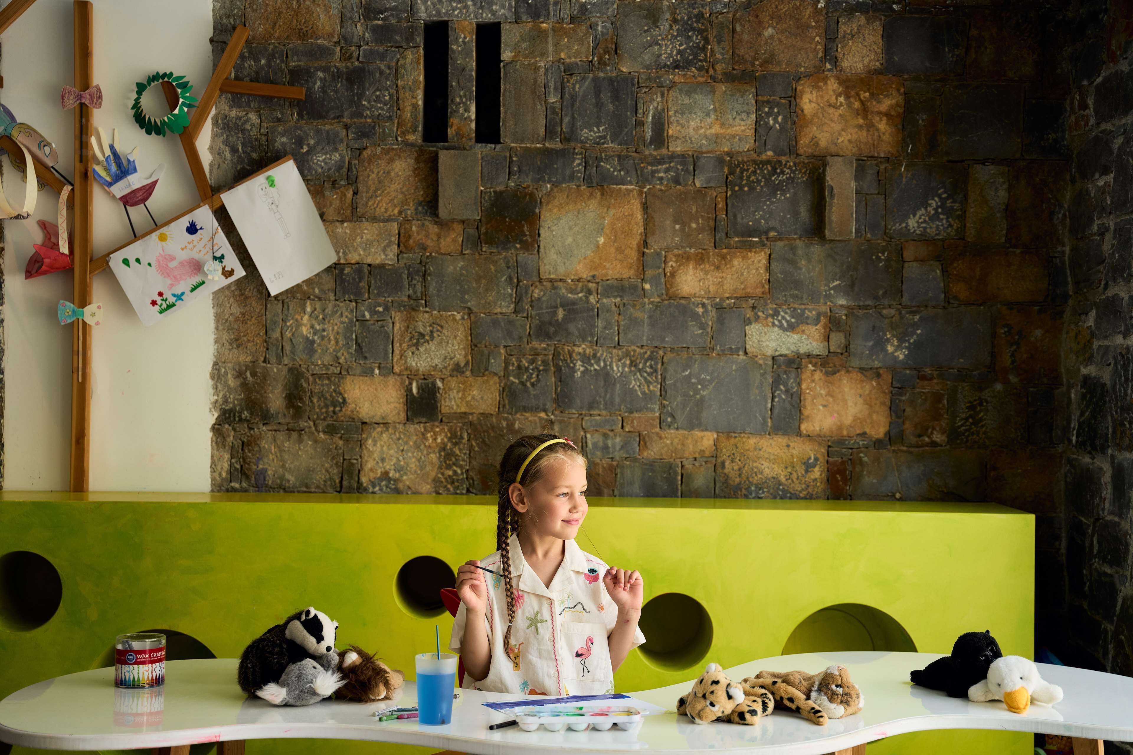 A child painting at a table with a stone wall background, a green bench, and various stuffed animals.