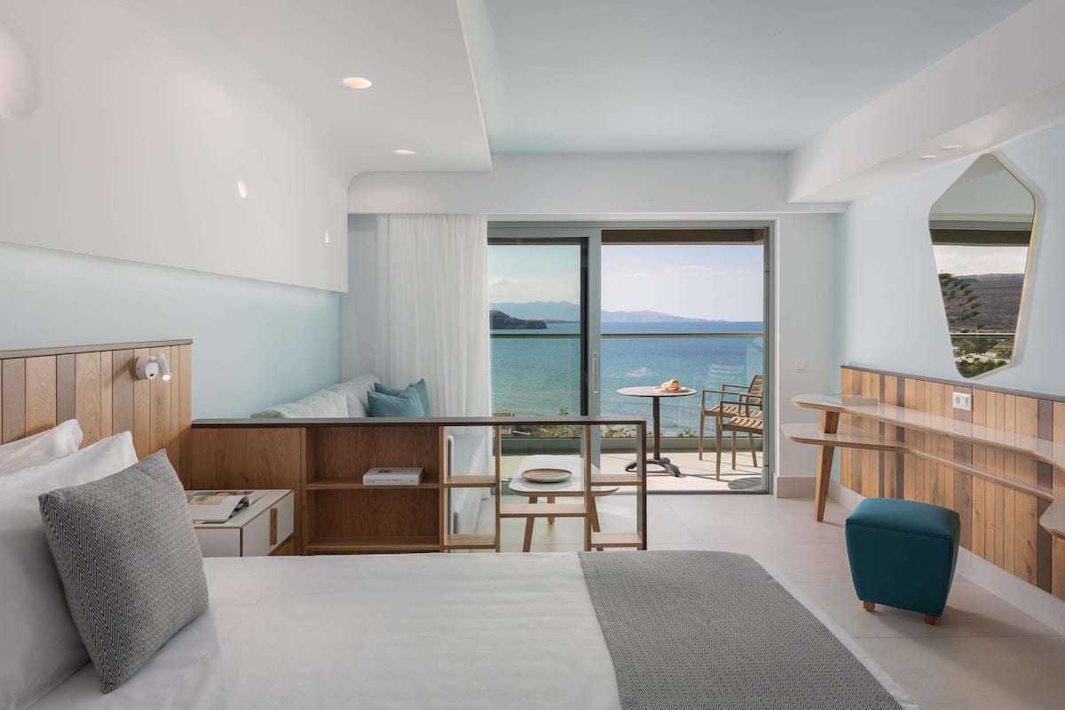 A modern hotel room with a large bed, a seating area with a view of the sea, and a balcony with a table and chairs.