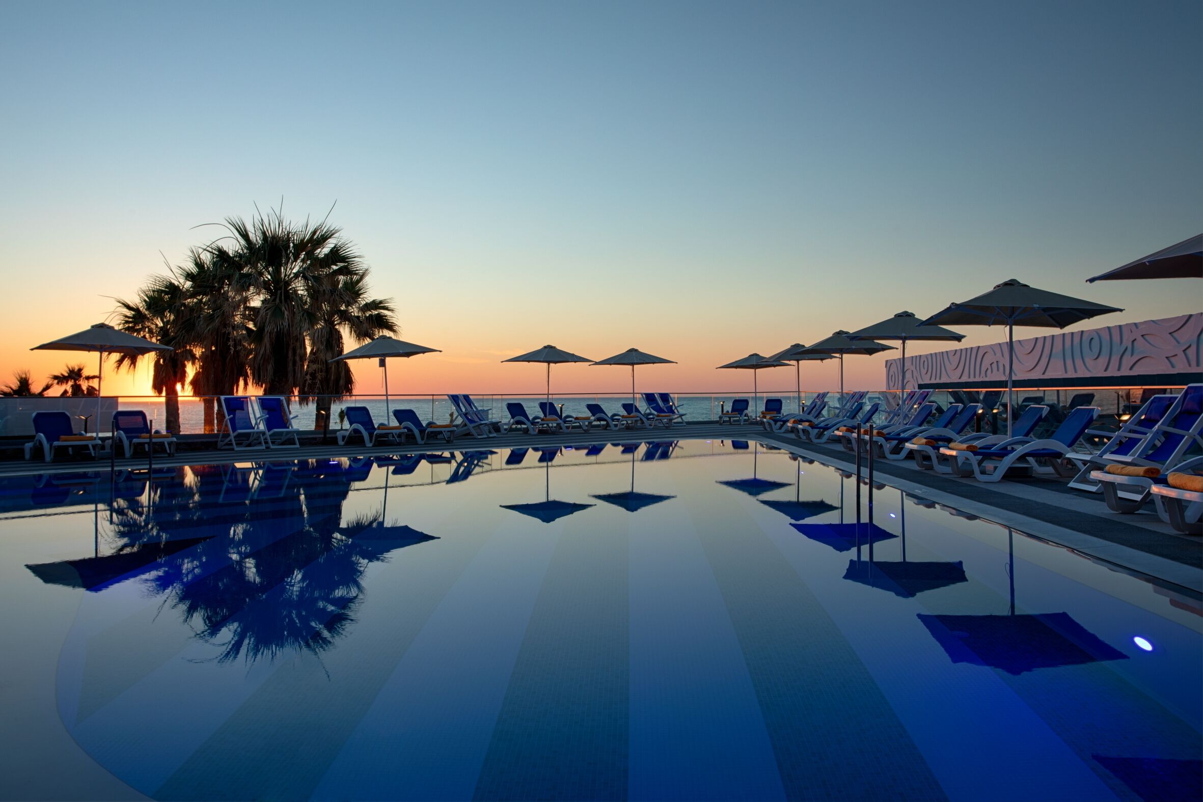 Ein Poolbereich mit Liegestühlen und Sonnenschirmen, Palmen und Blick auf einen Sonnenuntergang.