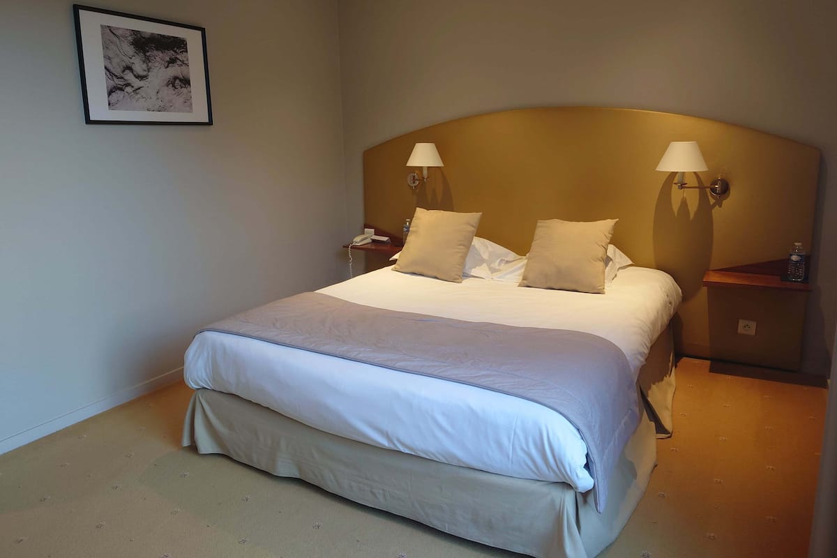 A hotel room with a bed, two bedside lamps, a nightstand with a phone, a framed picture on the wall, and a carpeted floor.
