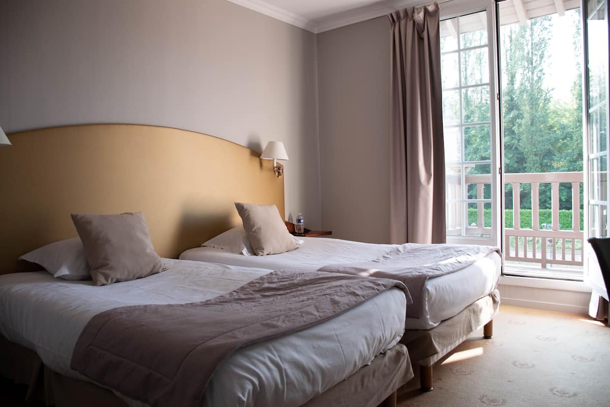 A bedroom with two beds, a nightstand, a lamp, and a view of the outdoors.