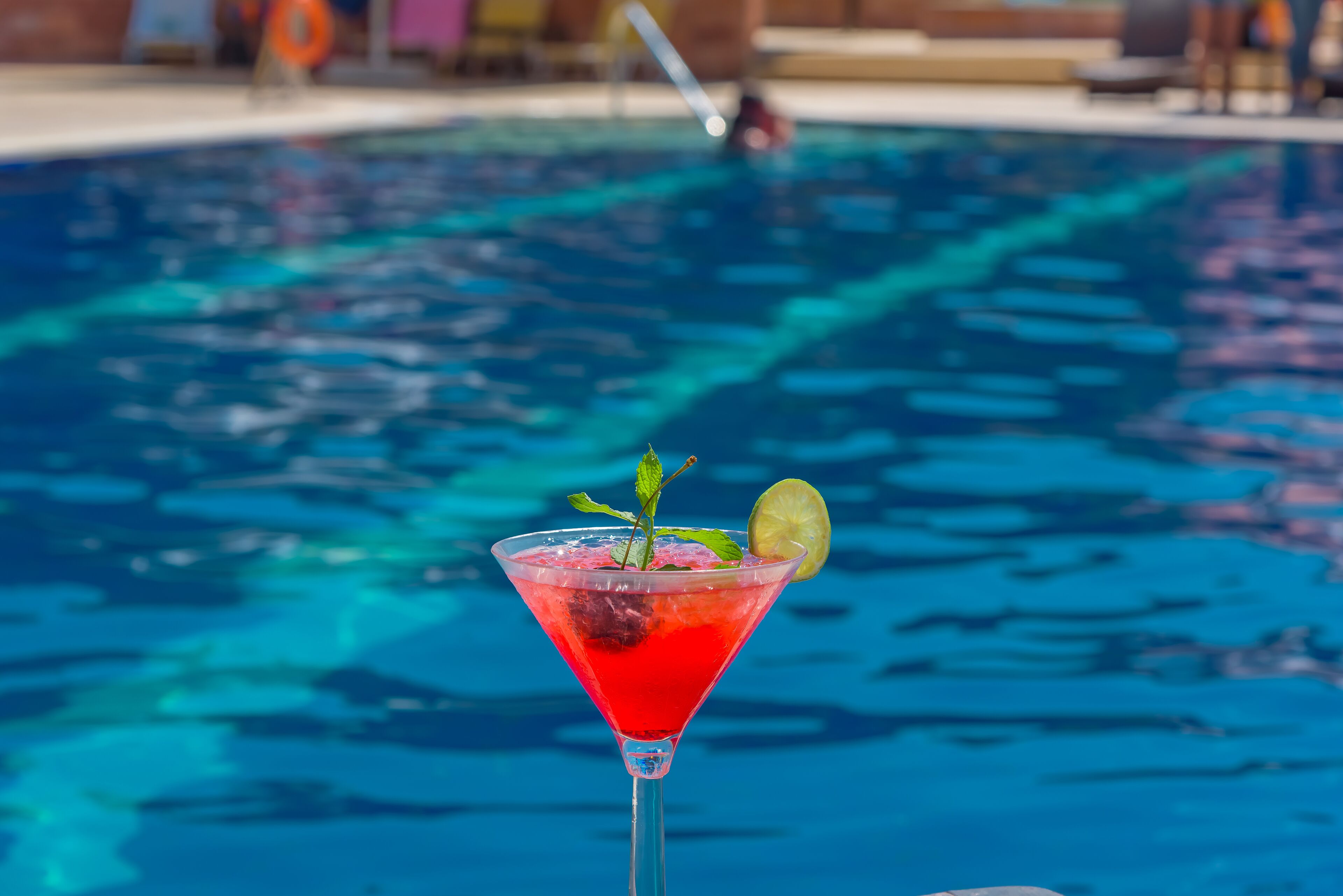 Ein freier Blick auf ein Schwimmbecken mit einem roten Cocktail in einem Martini-Glas, garniert mit Minze und einer Limettenscheibe.