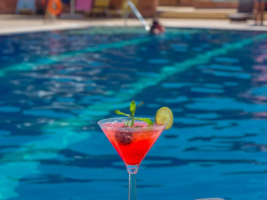 Vista clara de una piscina con un cóctel rojo en copa de martini, adornado con una hoja de menta y una rodaja de lima.