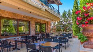 An outdoor dining area with multiple tables and chairs, large windows, and a view of the surrounding greenery. - Avalon Airport Hotel Thessaloniki (Thermi)