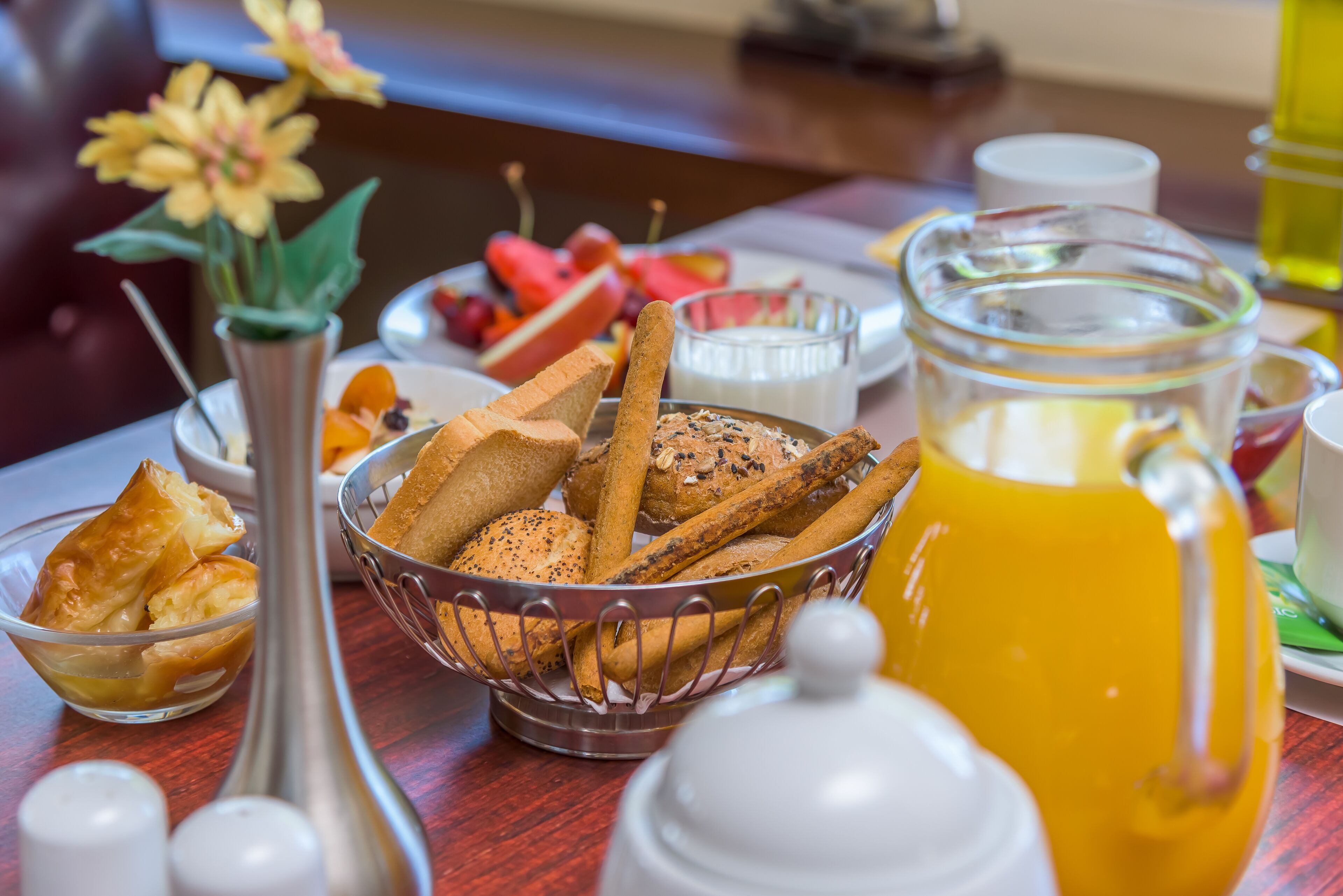 Una mesa de desayuno con pan, frutas, una jarra de jugo de naranja y un florero con flores.