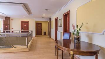 A hotel lobby with a wooden floor, a dining table with chairs, a glass railing, and a vase with flowers.