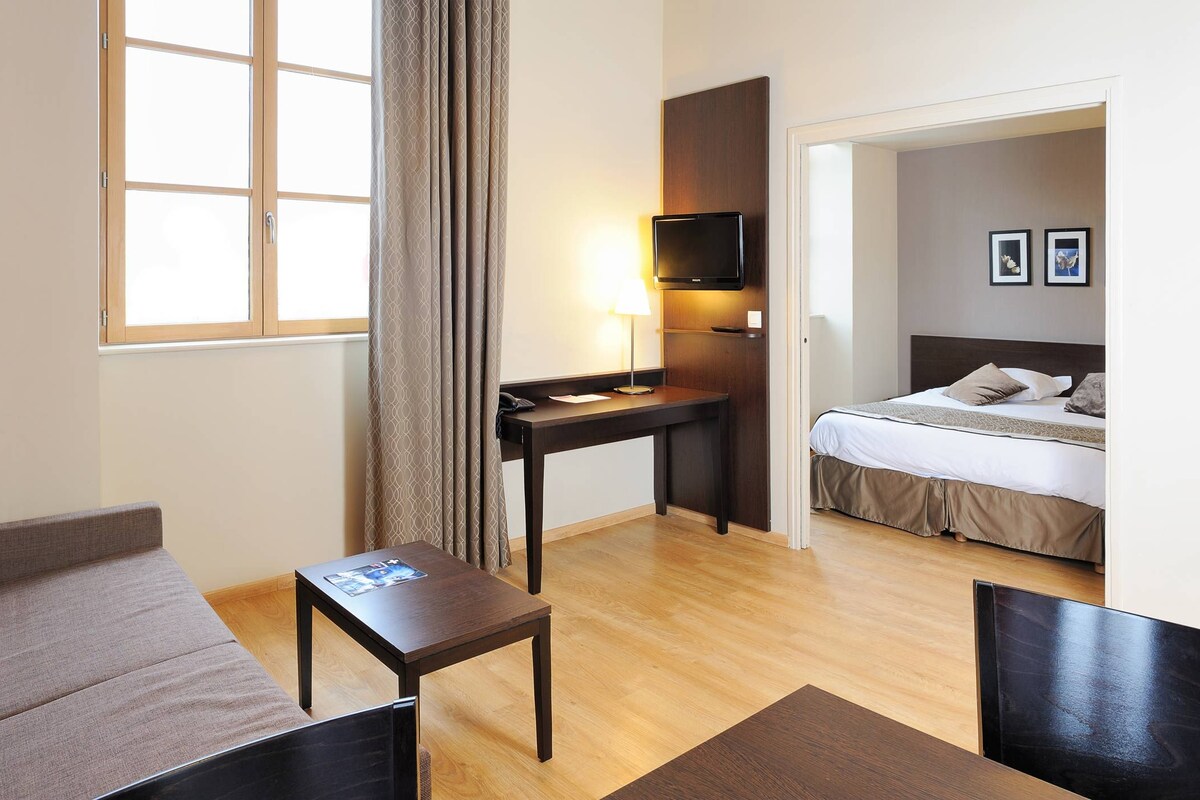 Habitación de hotel con una cama, un escritorio con lámpara, un televisor y una ventana con cortinas.