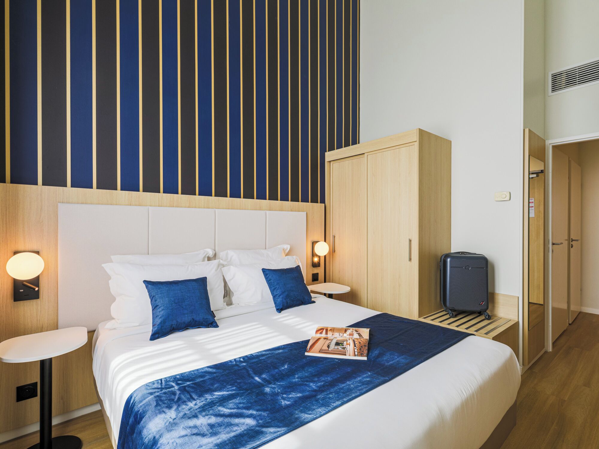 Een hotelkamer met een bed, nachtkastjes, een kledingkast, een koffer en een gestreept behang.