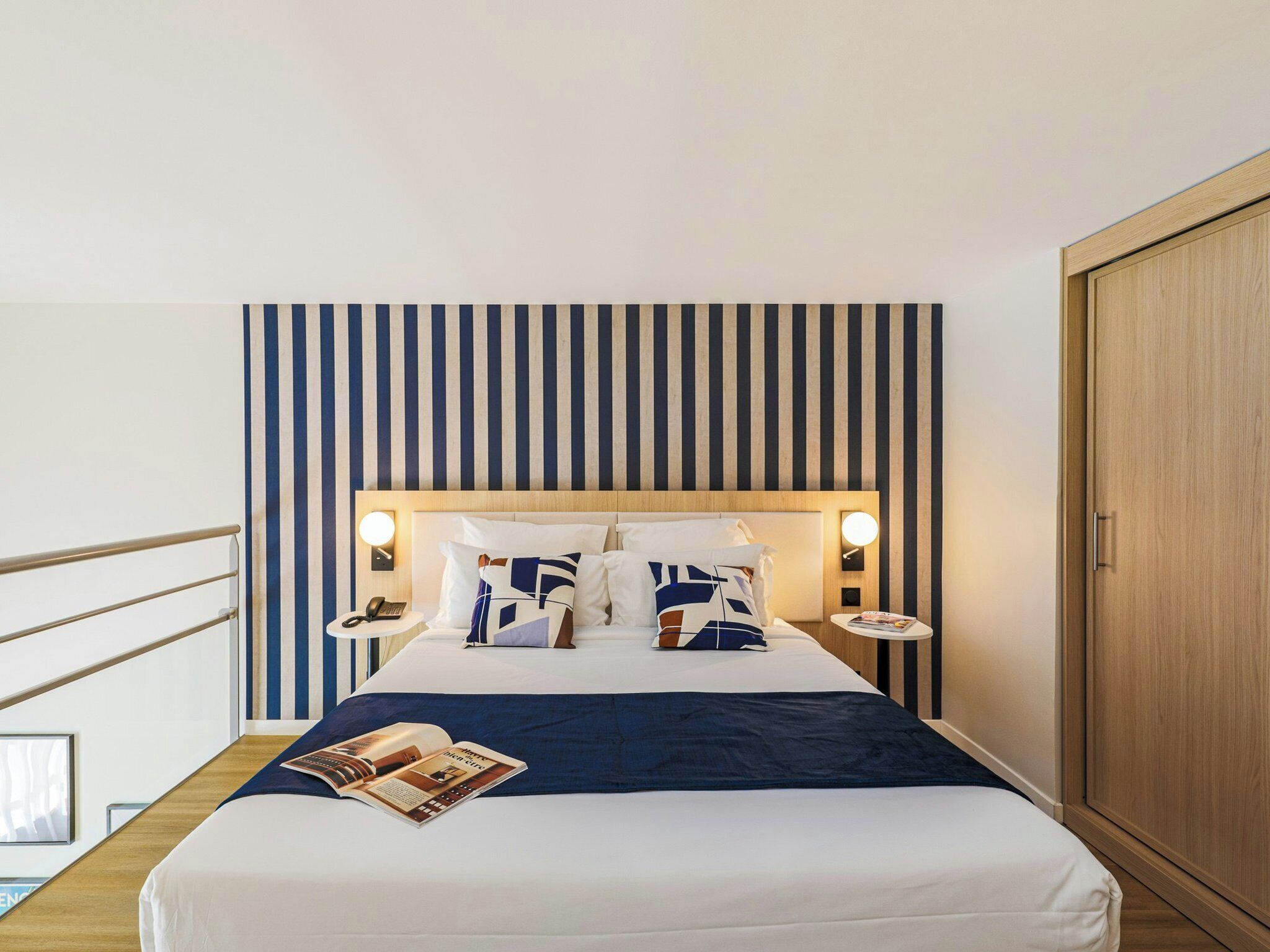 Hotelkamer met een bed, nachtkastjes, een tijdschrift, een houten kledingkast en een gestreepte wand.