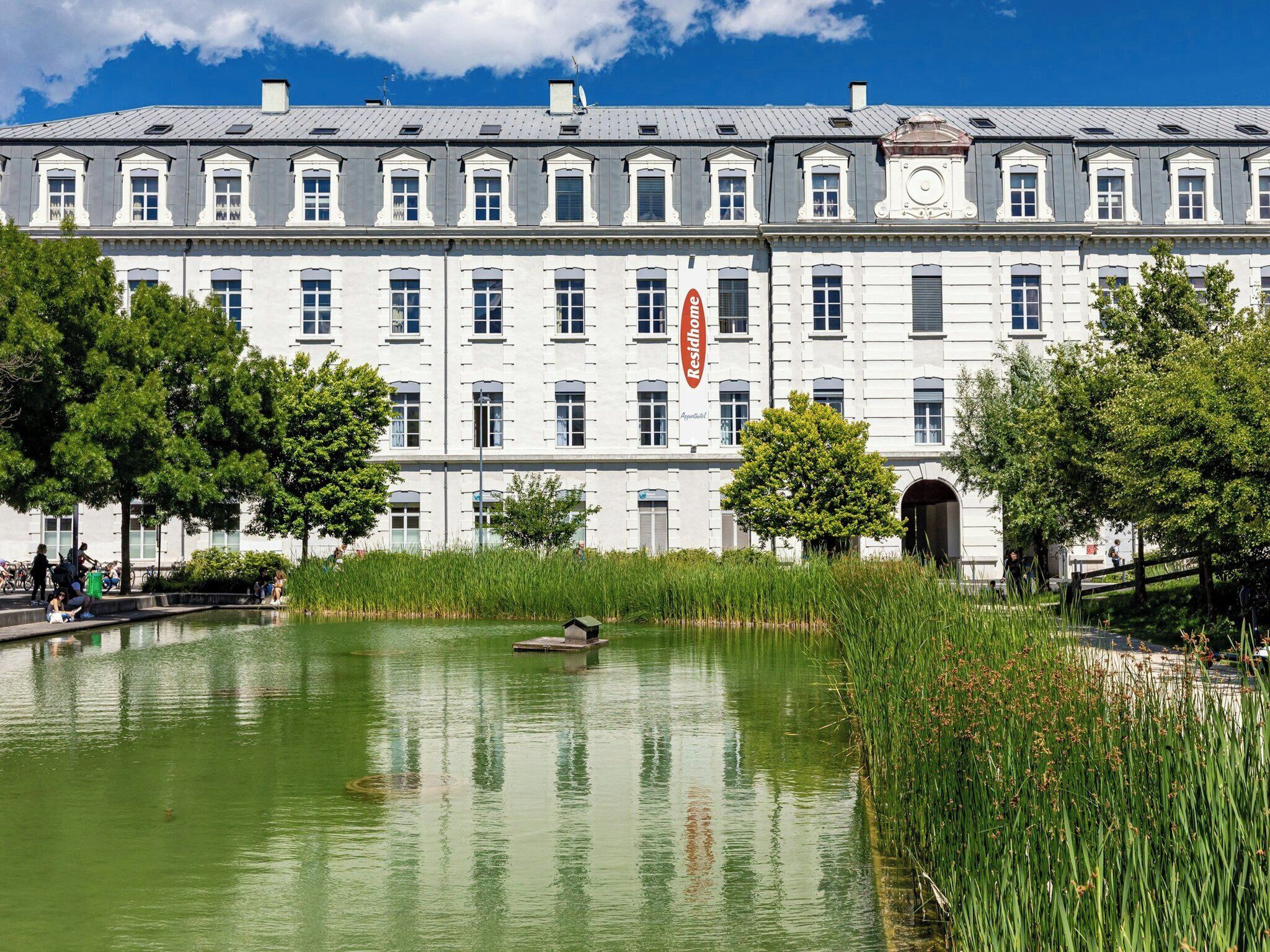 A large building with a pond in front, surrounded by trees and greenery.