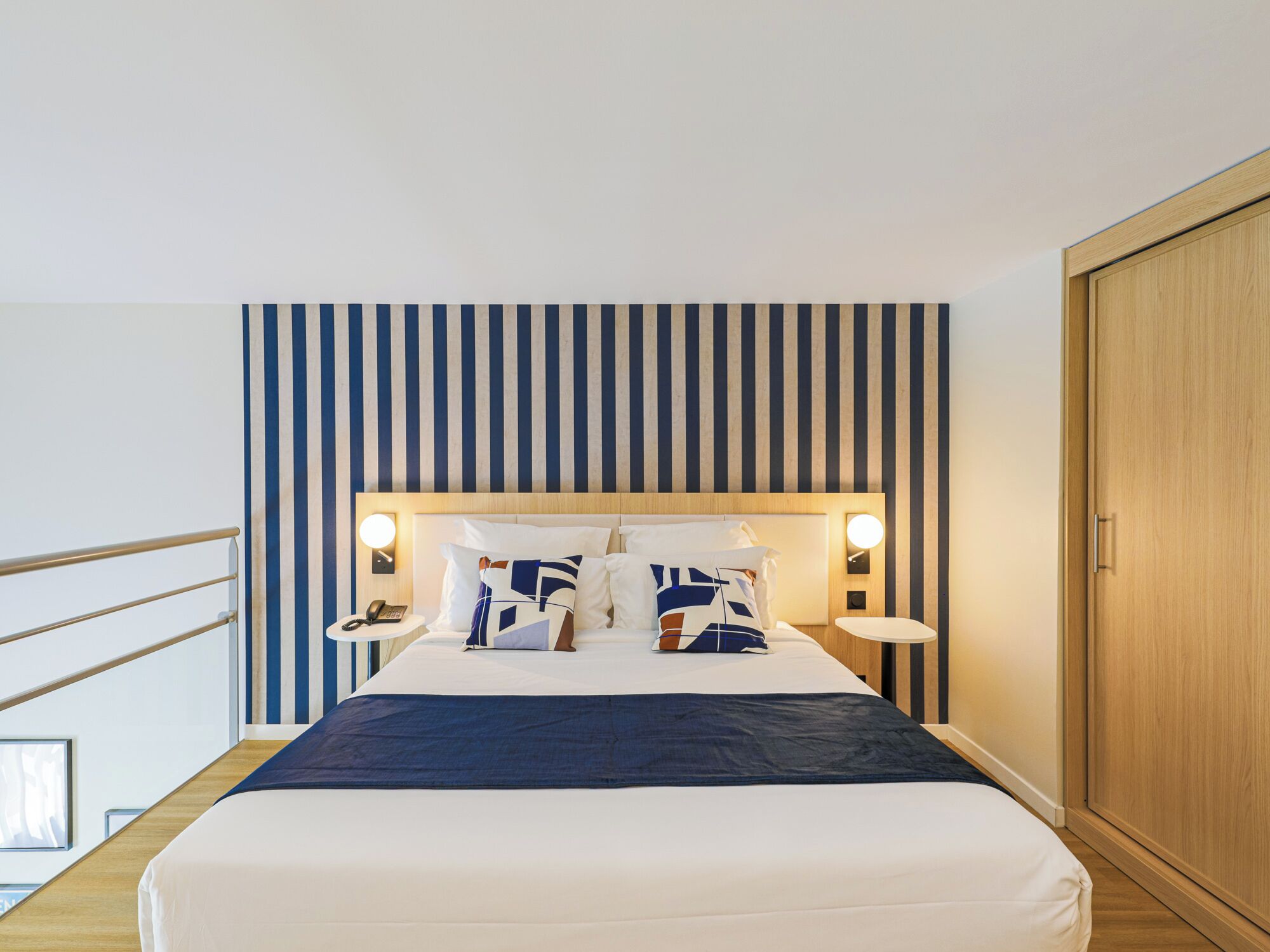 A hotel room with a bed, bedside tables, a wooden wardrobe, and a striped wall.