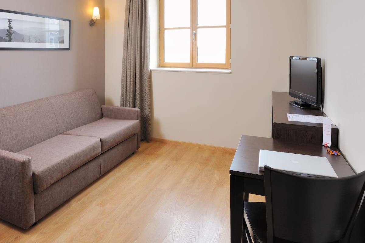 Een kamer met een bank, een bureau met een computer, een televisie en een raam met gordijnen.