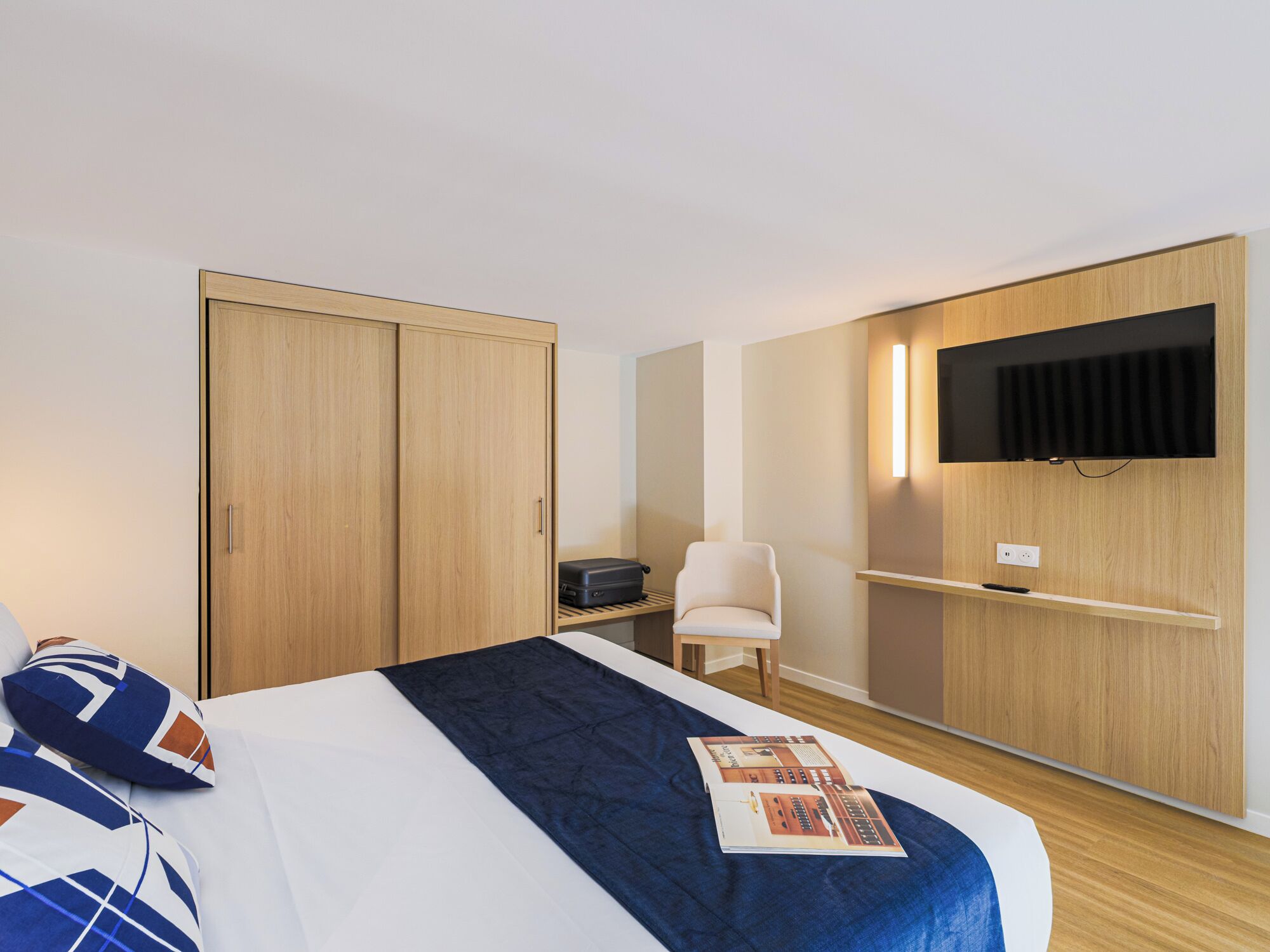 Una habitación de hotel con cama, una silla, un armario, un escritorio con televisor de pantalla plana y una maleta.
