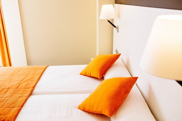 Una habitación de hotel con una cama, dos almohadas naranjas, una colcha blanca y una lámpara en la mesita de noche.