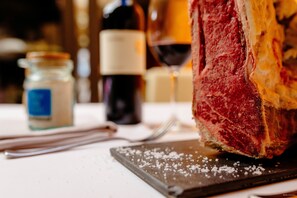 A large piece of meat on a cutting board with salt, a knife, and a wine bottle in the background. - Hotel Can Batiste (Sant Carles de la Rapita)