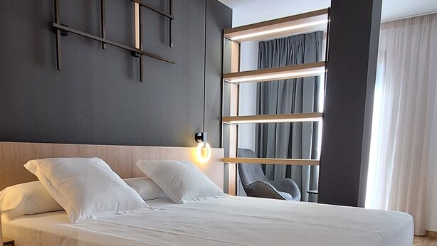 Moderni hotellihuone, jossa on suuri sänky, puinen pääty, seinään kiinnitetty valaisin ja ikkunasta avautuva näkymä kaupunkiin, ikkunassa on verhot.