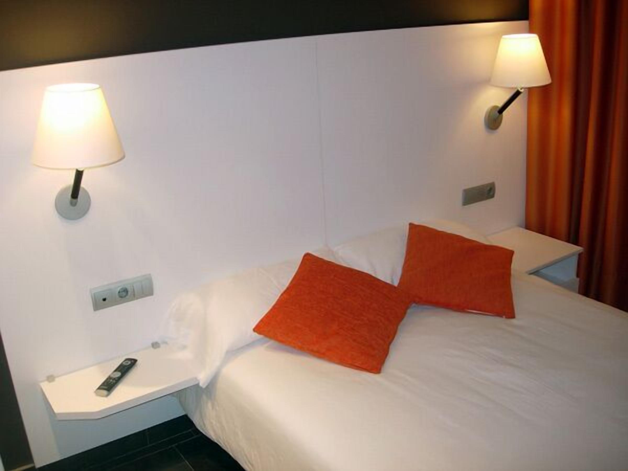 A hotel room with a bed, two nightstands, a remote control, and two wall-mounted lamps.