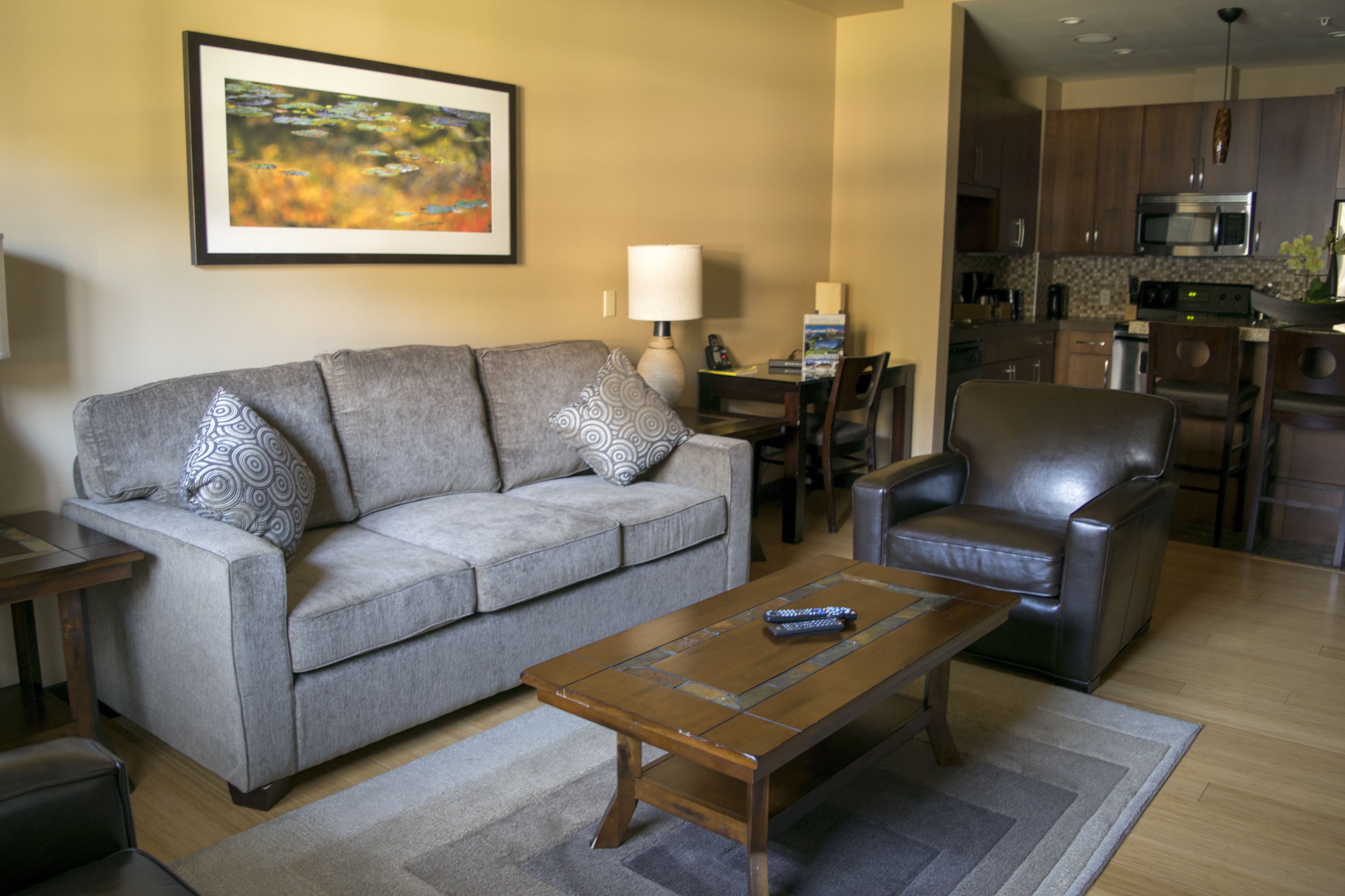Sala de estar com sofá cinzento, mesa de centro marrom e poltrona de couro marrom. Há um quadro na parede e uma luminária em uma mesinha lateral.