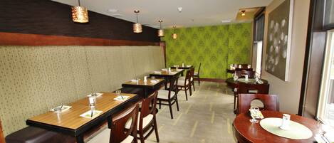A restaurant with multiple wooden tables and chairs, a patterned green wall, and pendant lighting.