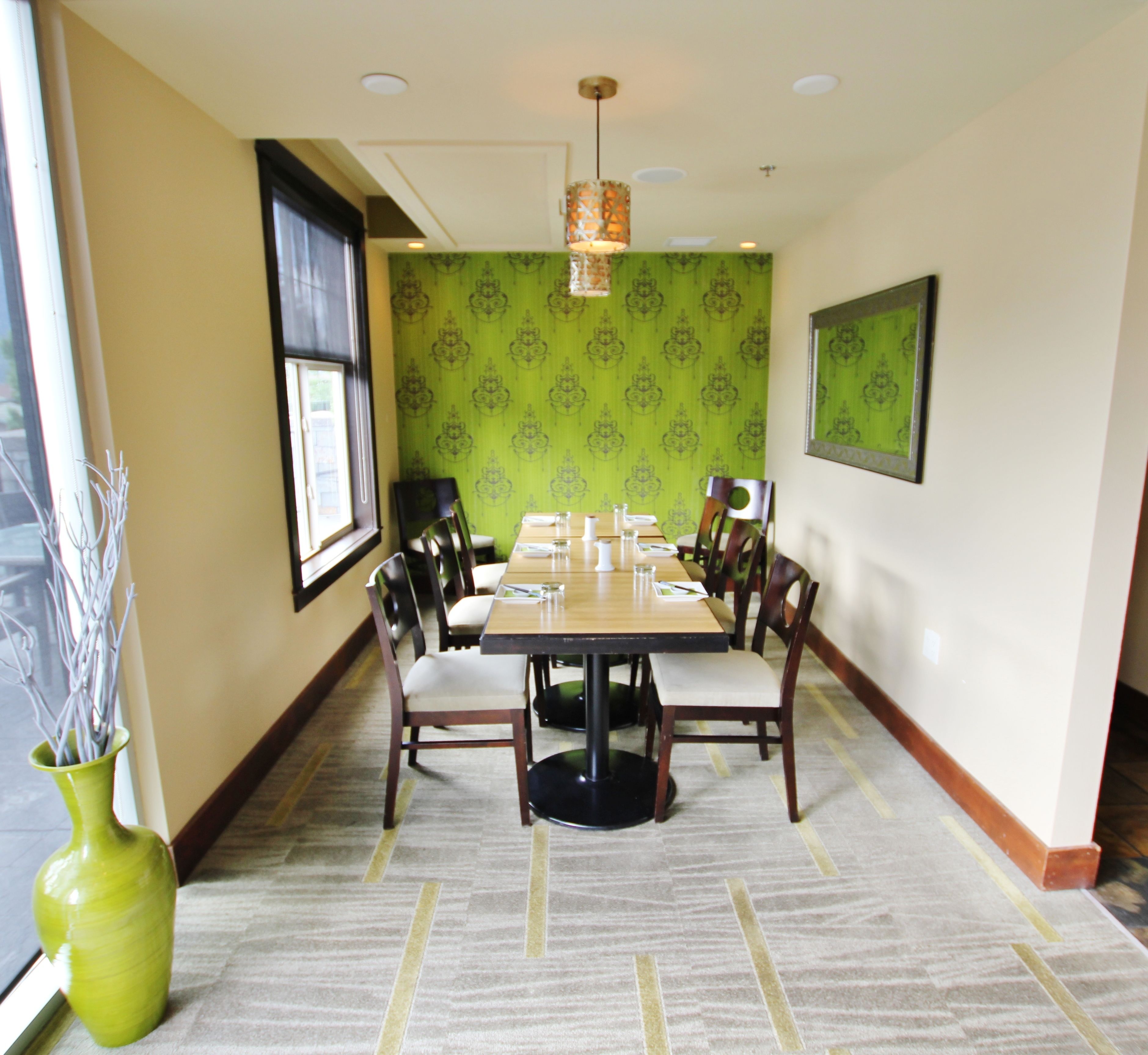 A dining area with a long table, chairs, a vase with branches, and patterned wallpaper.