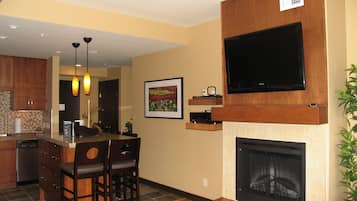Una sala de estar moderna con chimenea, un televisor de pantalla plana montado en una pared de madera, una zona de comedor con barra y una cocina con armarios de madera y lavavajillas.