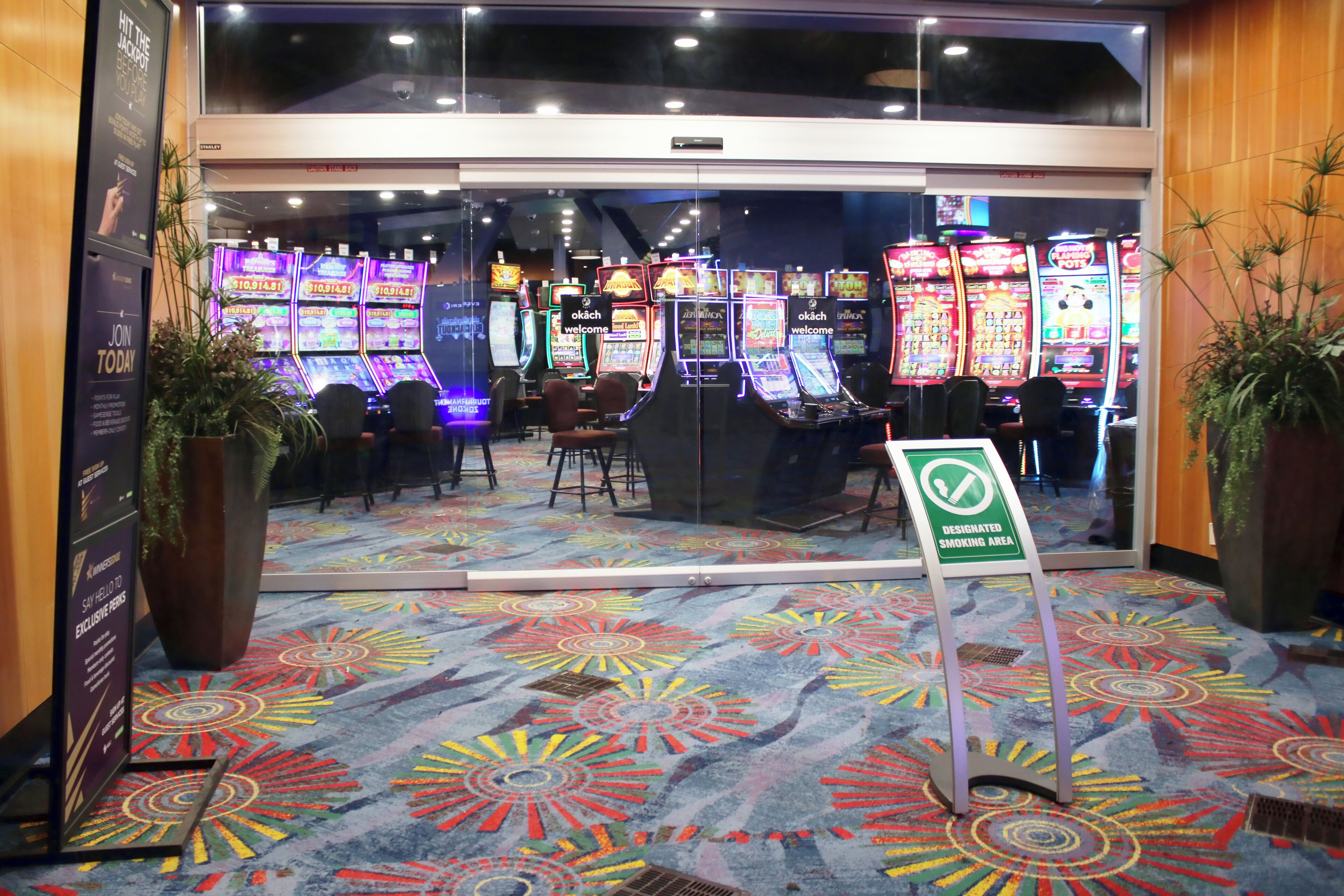 L’entrée d’un casino, avec des machines à sous, un panneau indiquant l’emplacement réservé aux fumeurs et une moquette aux couleurs vives.