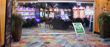 L’entrée d’un casino, avec des machines à sous, un panneau indiquant l’emplacement réservé aux fumeurs et une moquette aux couleurs vives.