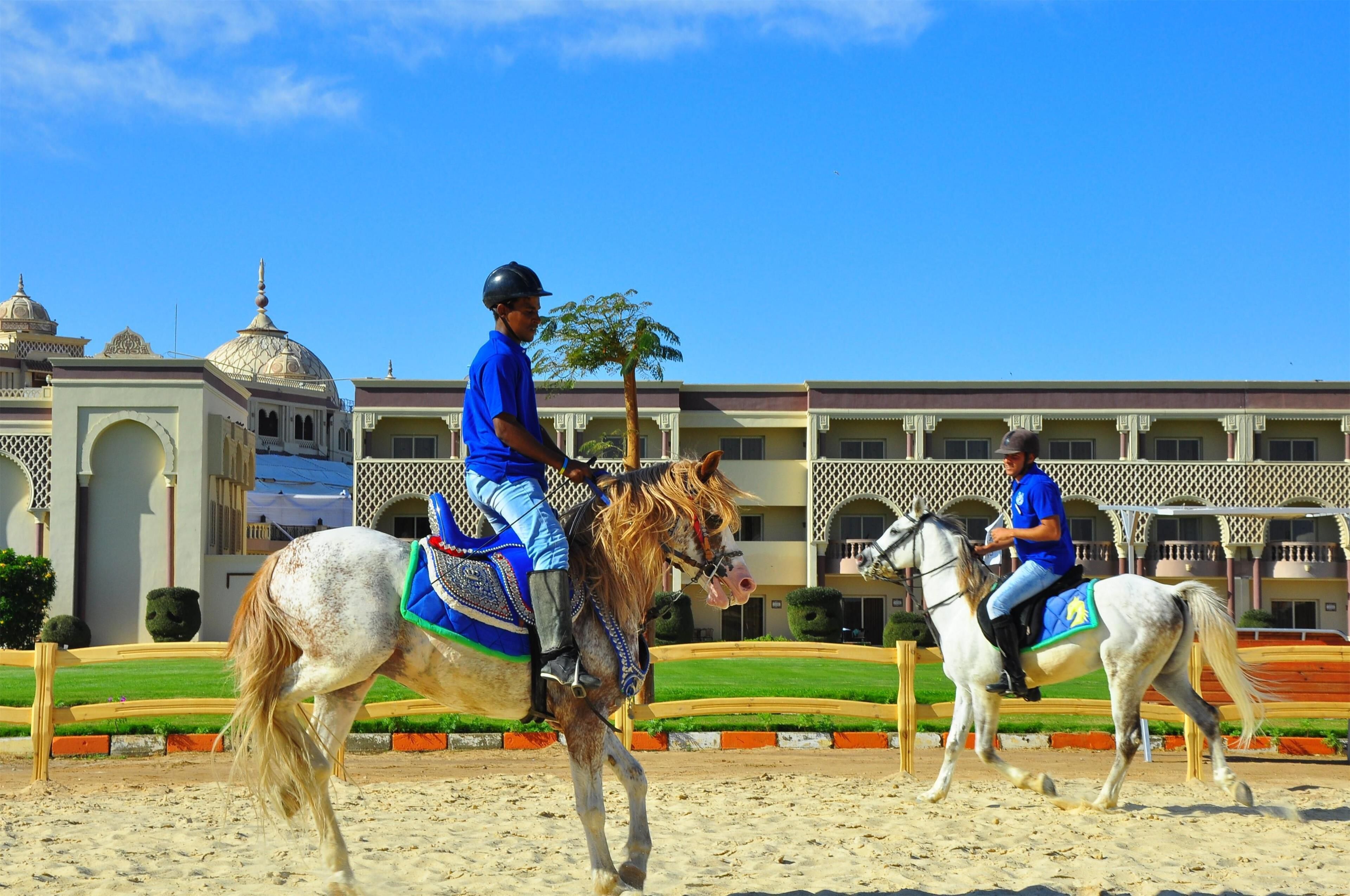 Dos jinetes a caballo en una arena, con un edificio al fondo.