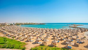 A beach with numerous sun umbrellas and lounge chairs, a clear blue sea, and a pier extending into the water. - Sunrise Sentido Mamlouk Palace - All inclusive (Hurghada)