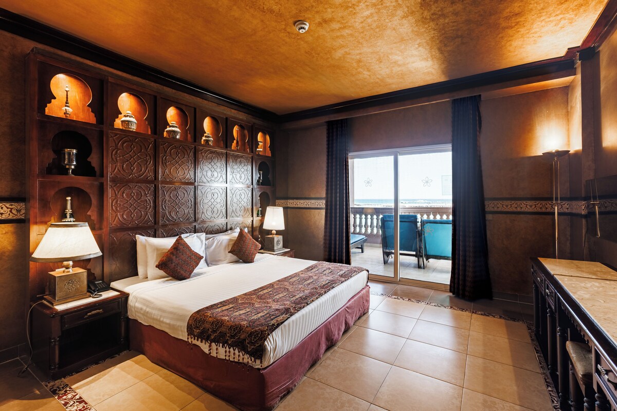 A hotel room with a large bed, a desk, a view of the outdoors, and a decorative wall panel.