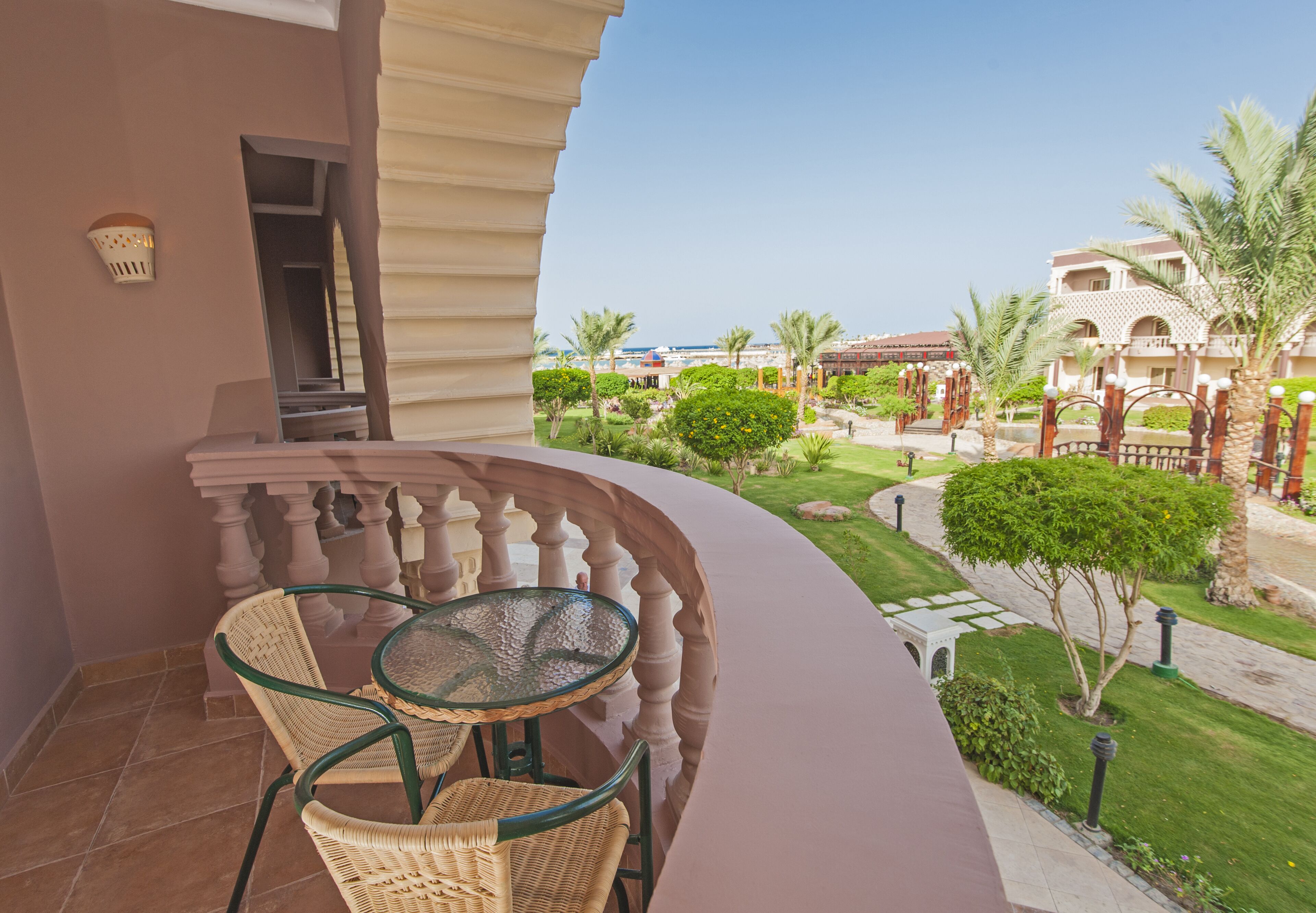 A balcony with a glass table and wicker chairs, overlooking a well-maintained garden with palm trees and a pathway.