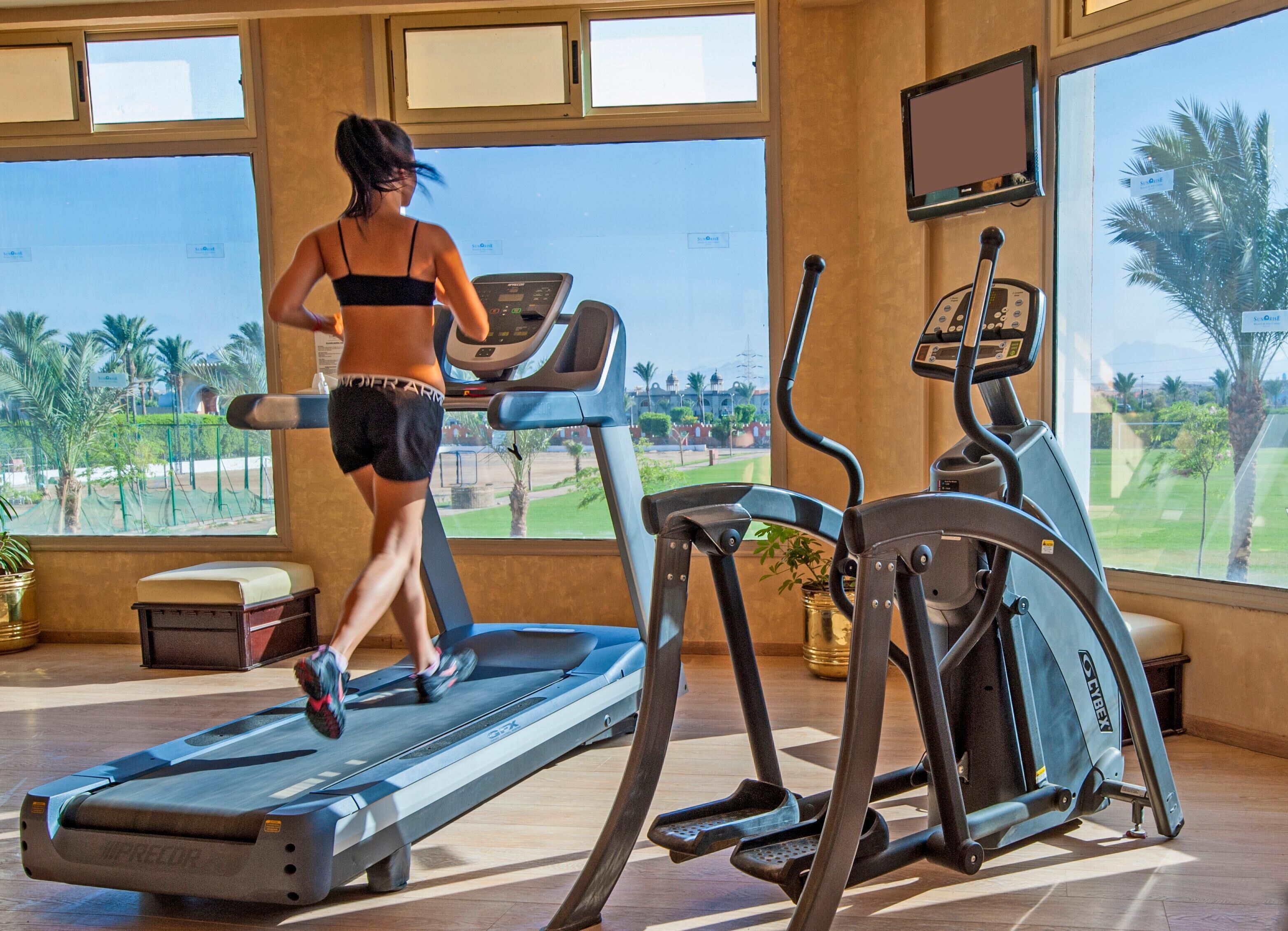 Una palestra con tapis roulant e attrezzature per esercizi, ampie finestre che offrono vista su palme e un campo da golf.