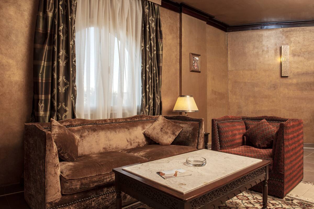 Una habitación con un sofá marrón, un sillón estampado, una mesa con un vaso y una lámpara.