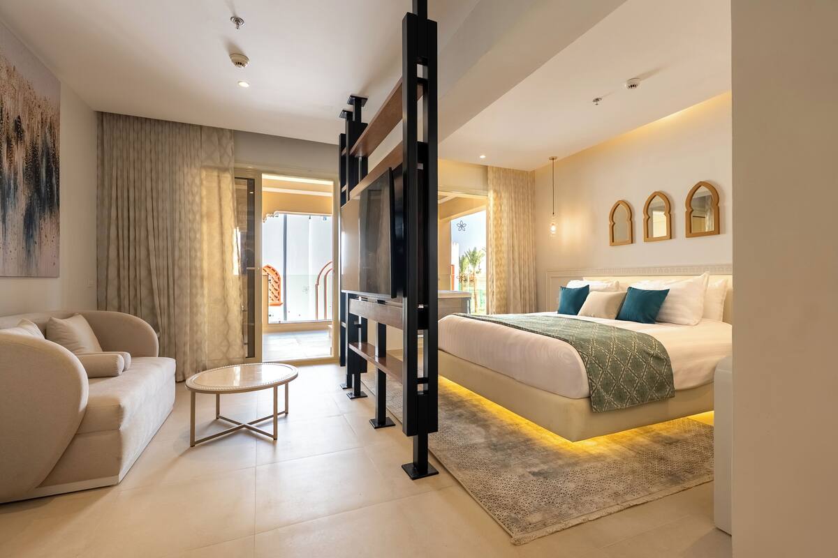 Una habitación de hotel moderna con sofá, una mesa redonda de centro, una cama con manta verde y un televisor grande.